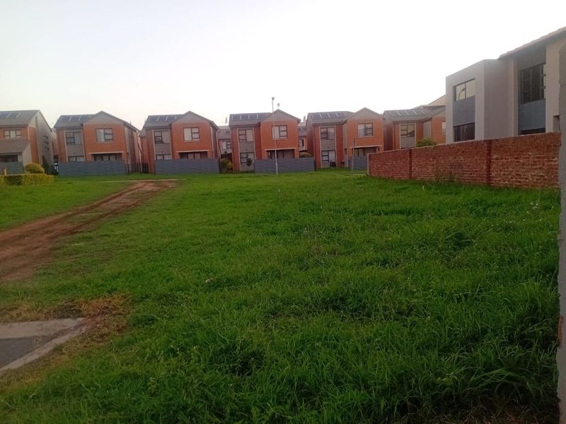 A vacant residential plot featuring a grass lawn and brick boundary wall, situated in a developed neighborhood with surrounding residential homes.