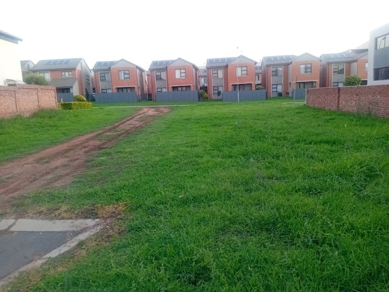 A vacant residential plot featuring a grass lawn, dirt driveway, and brick boundary walls, situated within a developed residential estate.