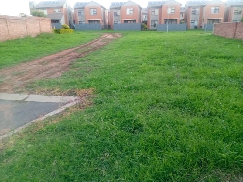 A vacant residential plot featuring grass coverage, brick boundary walls, and established driveway access within a developed residential estate.