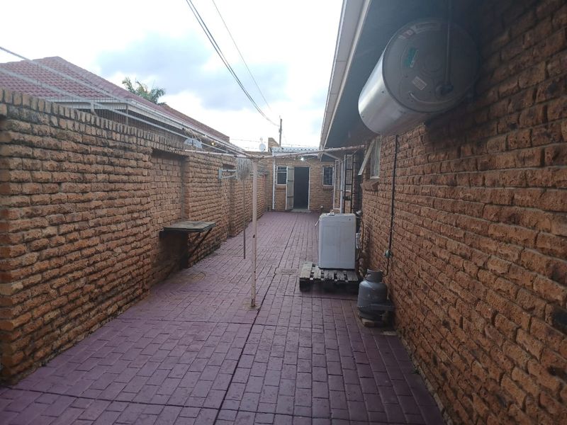 A narrow rear exterior walkway featuring brick boundary walls, paved flooring, and an external geyser unit.