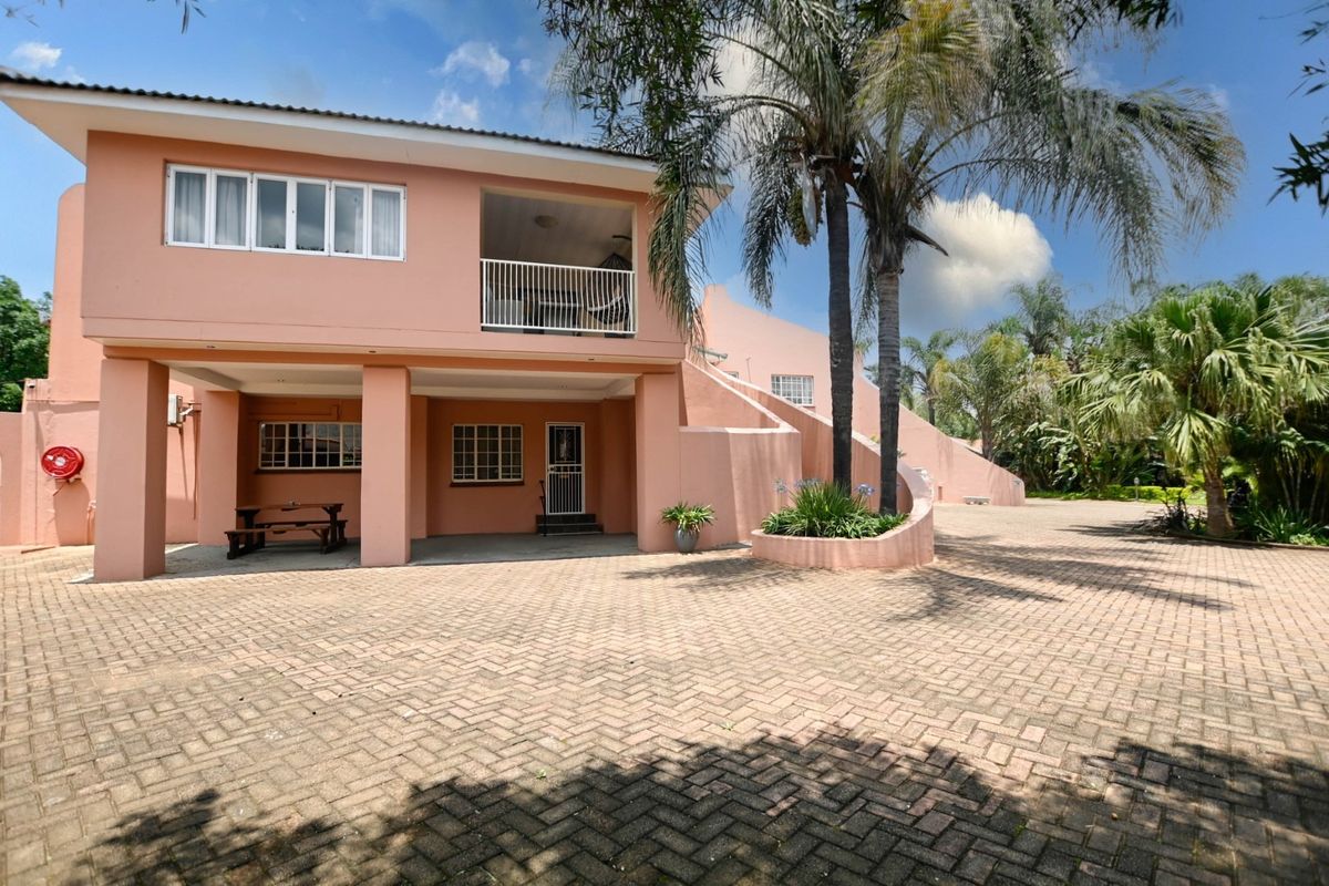 Rear view of the property featuring a large paved driveway, covered patio area, upper-level balcony, and established garden landscaping.