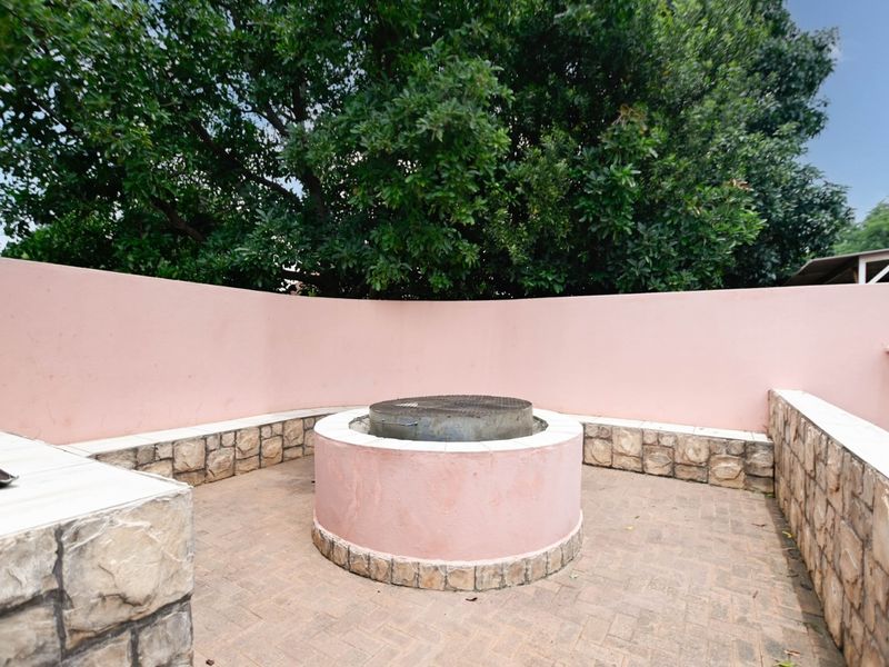 Outdoor entertainment area featuring a circular built-in braai, stone-clad retaining walls, and paved flooring, set against a backdrop of mature trees.