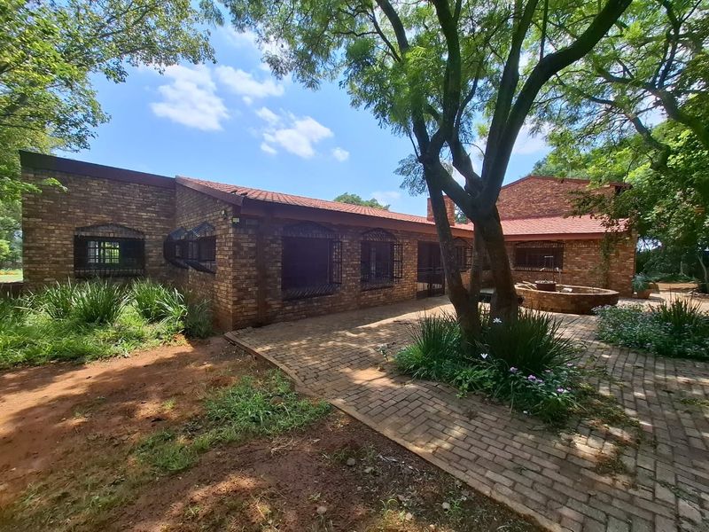 Rear view of a face brick farmhouse featuring a paved patio area, mature trees, and a stone water feature in a garden setting.