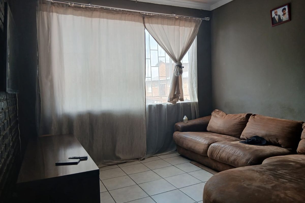 A living room featuring tiled flooring, an exposed brick wall, and a window fitted with security bars.