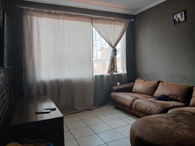 A living room featuring tiled flooring, an exposed brick wall, and a window fitted with security bars.