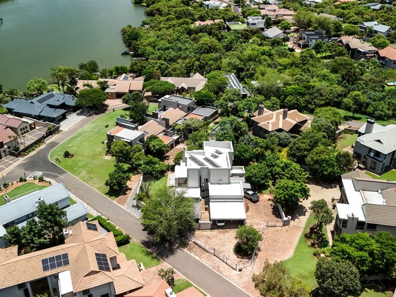 An aerial view of a residential estate featuring multiple homes, lush green landscaping, paved roads, and proximity to a large water body.