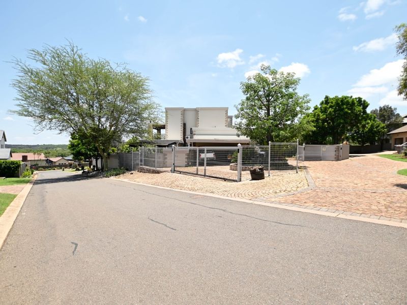 A street-level view of the property entrance, featuring a paved driveway, secure wire mesh fencing, and established trees in a residential estate setting.