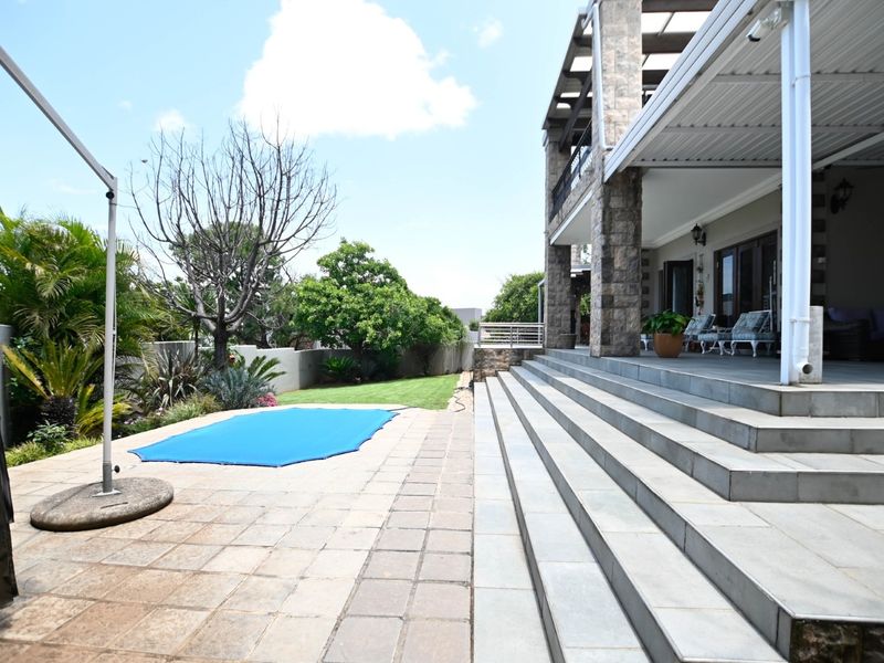 Spacious rear exterior featuring a covered patio with stone pillars, tiled steps leading to a paved pool area, and a well-maintained garden.