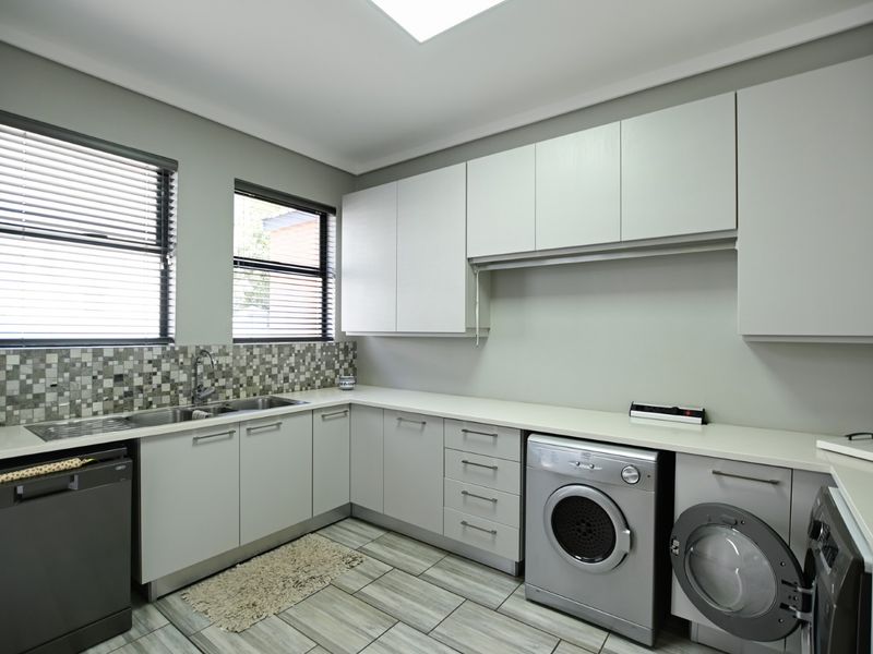 A modern, well-appointed laundry room featuring extensive built-in cabinetry, stone countertops, a double sink, mosaic backsplash, and a skylight for natural illumination.