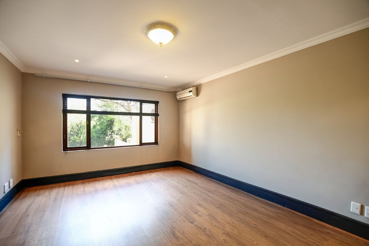 A bright, empty bedroom featuring laminate flooring, an air conditioning unit, and large windows with views of the surrounding greenery.