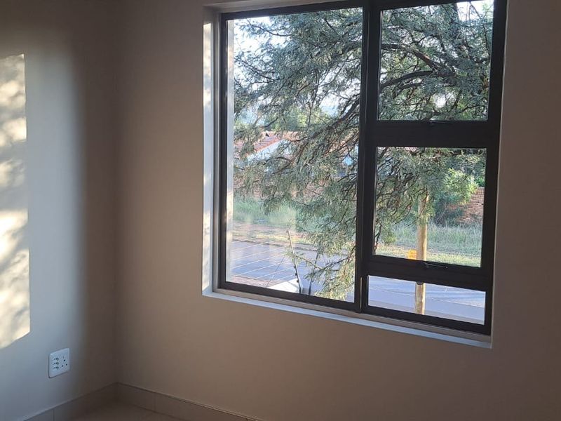 A bright, empty bedroom featuring neutral walls, tiled flooring, and a large aluminium-framed window with views of the surrounding area.