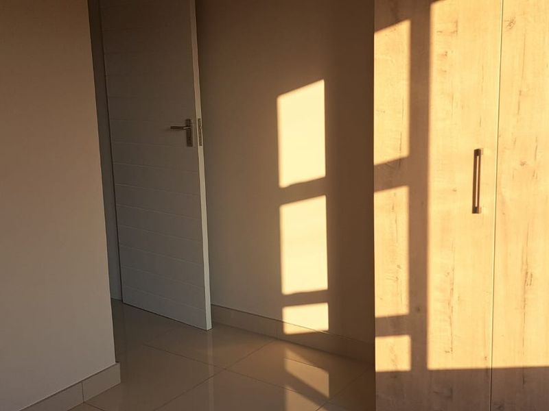 A bright, empty bedroom featuring modern light wood-finish built-in cupboards and neutral tiled flooring, illuminated by natural sunlight.