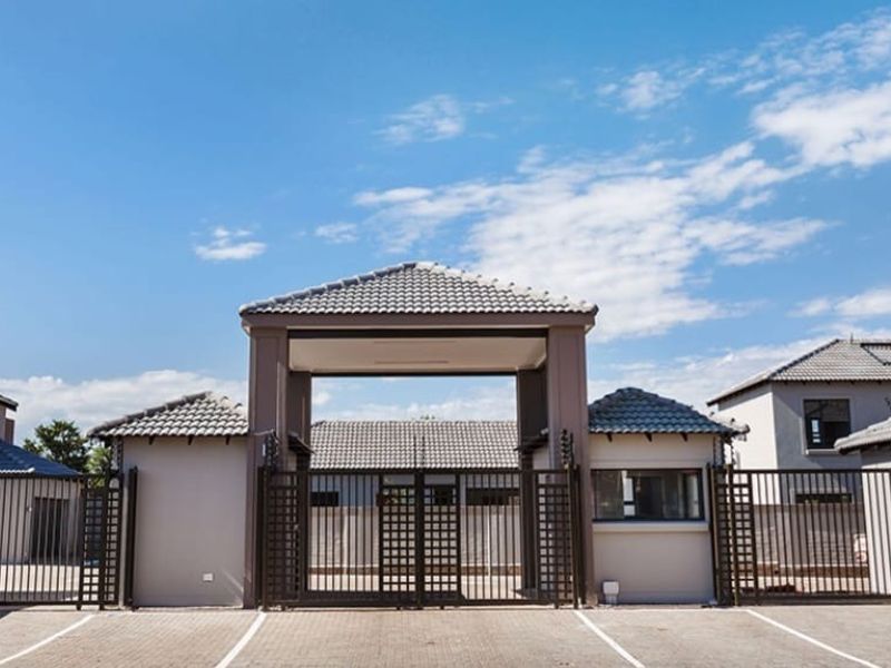The front entrance of the townhouse complex features a secure gated access point with a guard house, paved driveway, and perimeter electric fencing.