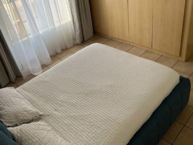 A bedroom featuring built-in wooden cupboards and tiled flooring, with natural light entering through a window with sheer curtains.