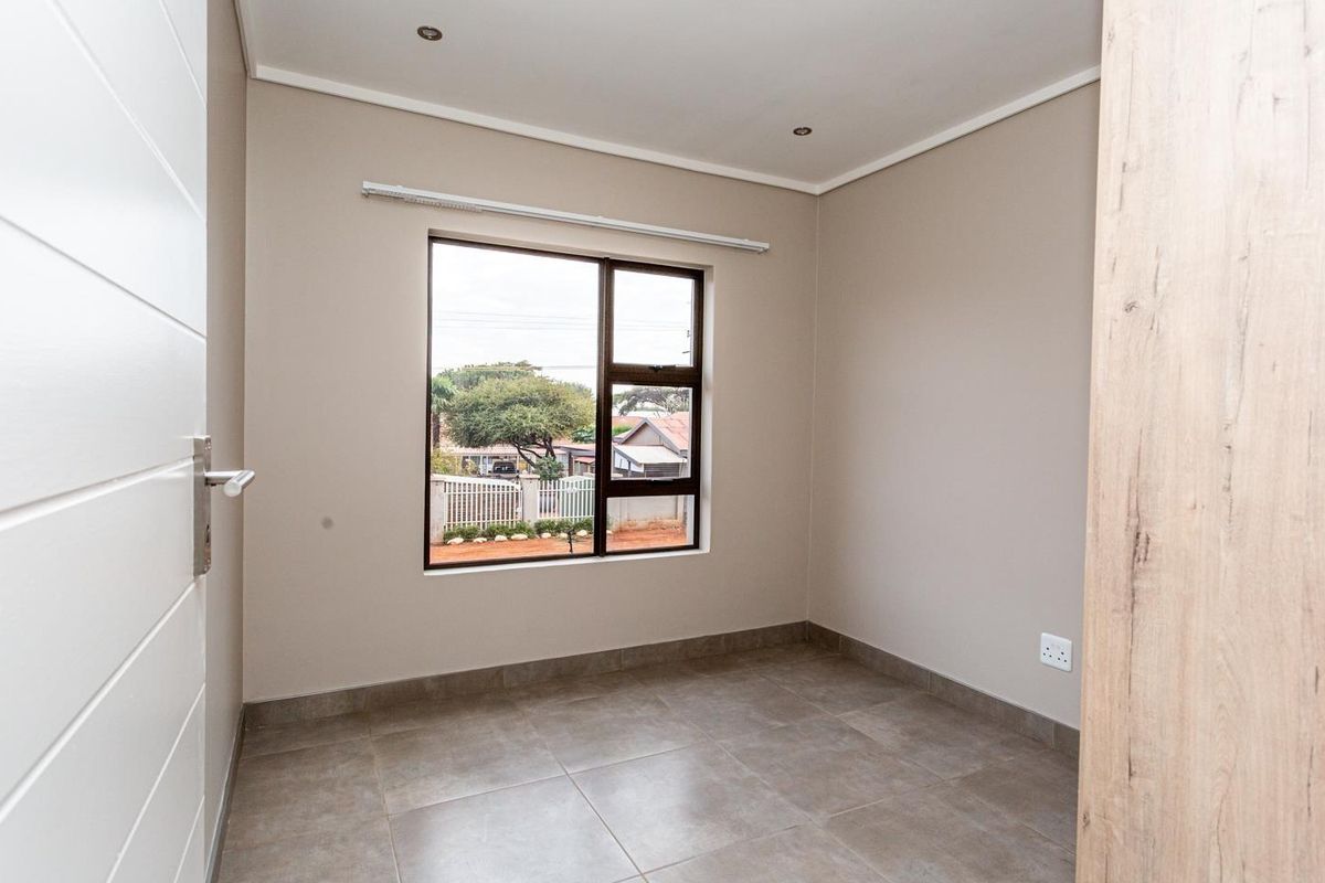 A clean, empty bedroom featuring neutral wall tones, tiled flooring, recessed downlights, and an aluminium-framed window providing natural light.