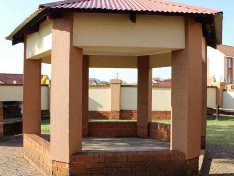 A detached outdoor gazebo featuring a corrugated metal roof, brick masonry base, and an adjacent built-in braai area within a paved garden setting.