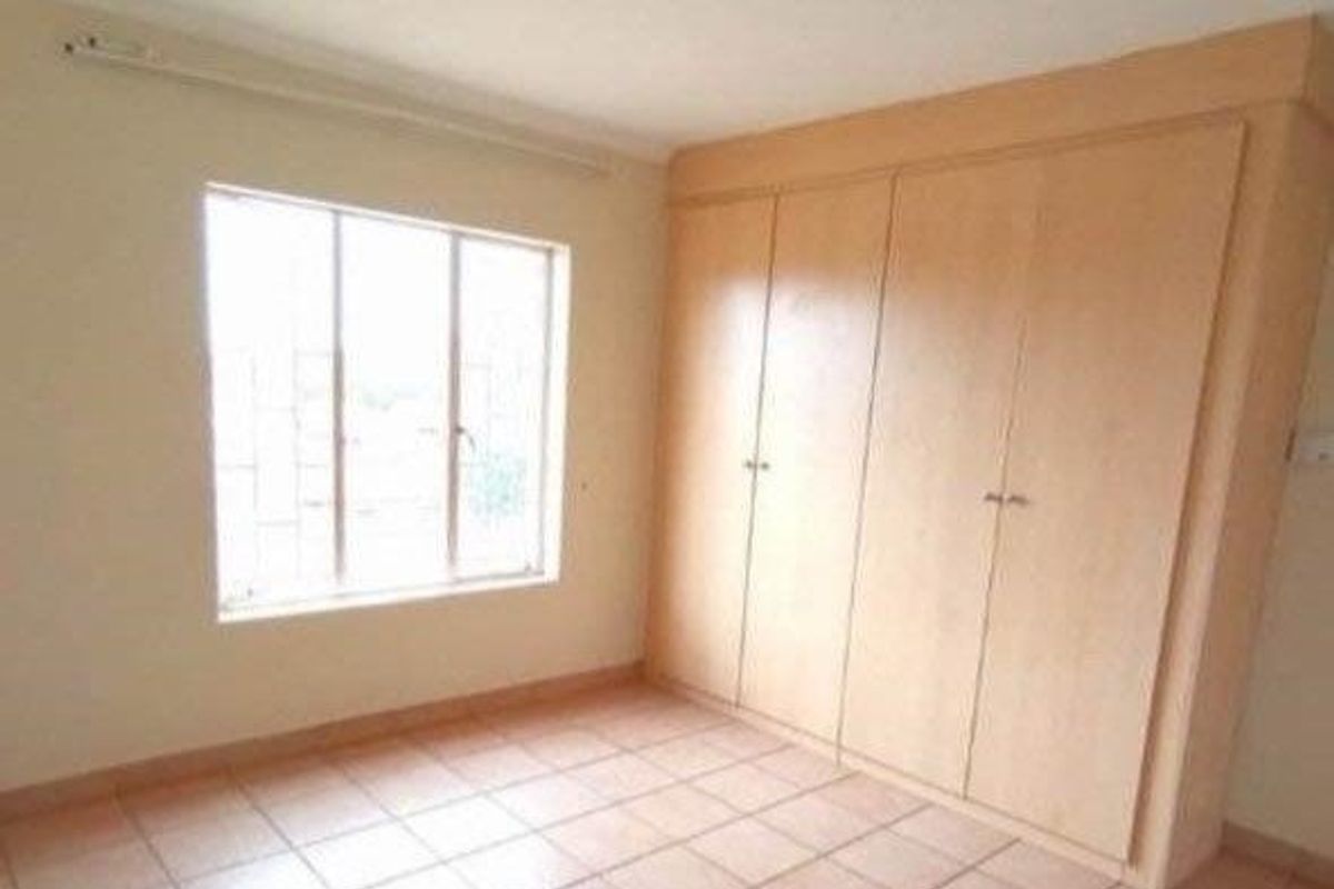 A bright, empty bedroom featuring tiled flooring, a large window with security bars, and ample built-in cupboard storage.