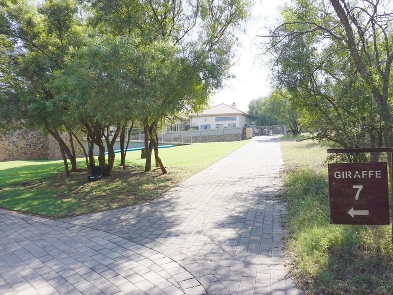 A paved driveway leading towards a residential property, surrounded by mature trees and well-maintained garden areas.