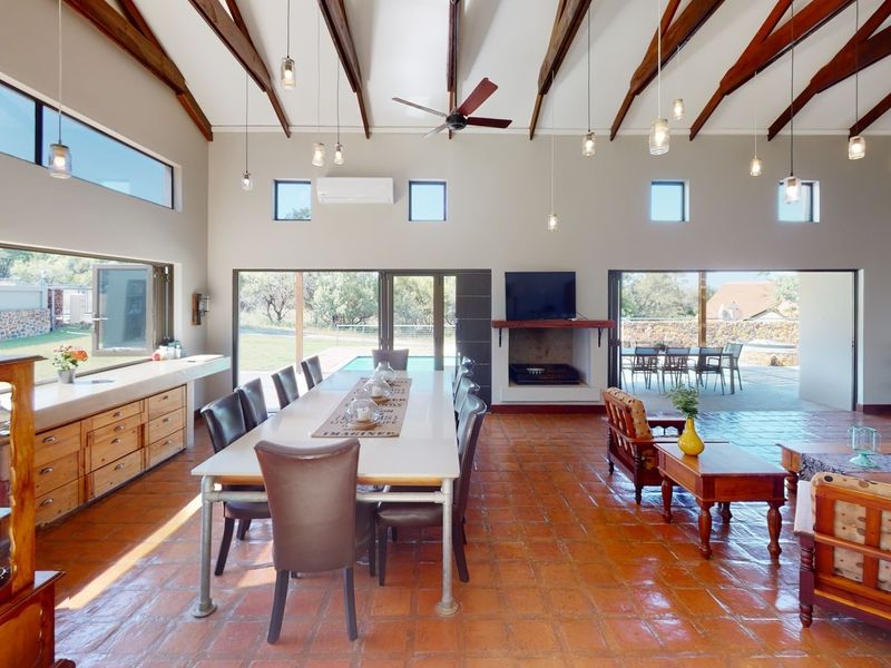 Spacious open-plan living and dining area featuring high vaulted ceilings with exposed timber beams, terracotta flooring, a built-in fireplace, and seamless access to the patio.