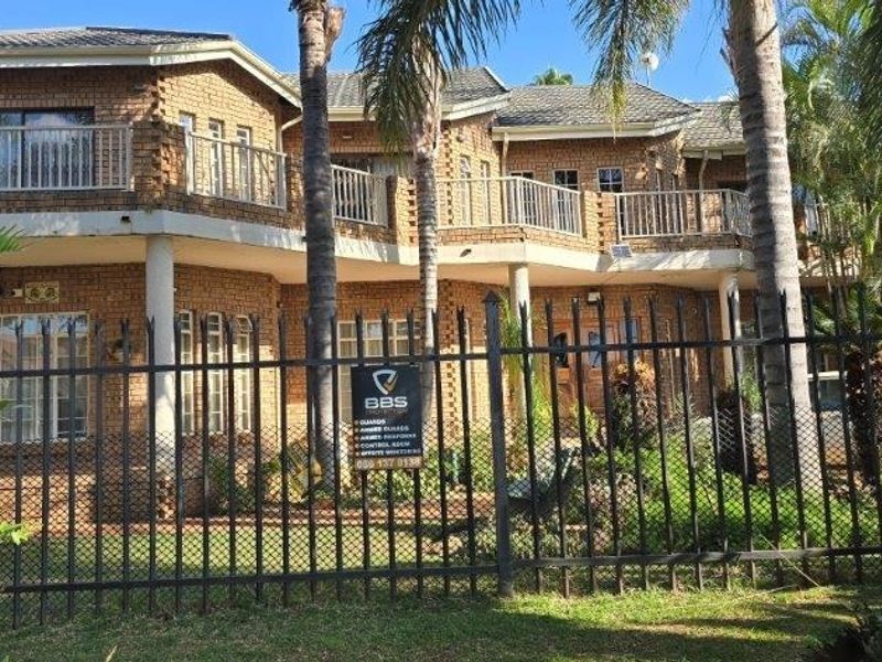 A two-story face brick residential building featuring upper-level balconies with metal railings, set behind secure perimeter fencing.
