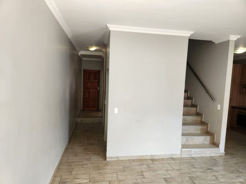 A bright, empty hallway featuring tiled flooring, a staircase with a metal handrail, and coved cornicing leading to a wooden entrance door.