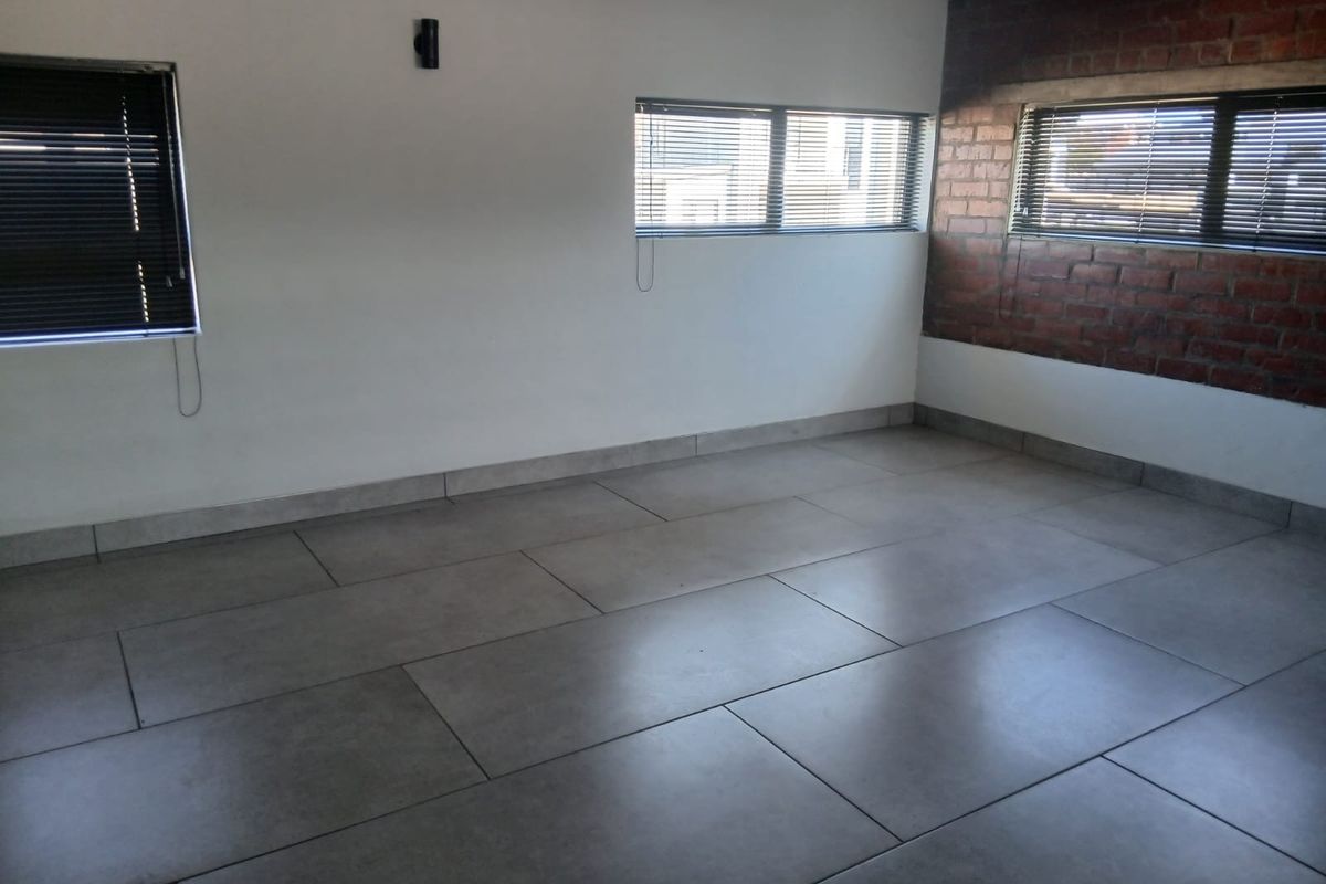 An empty living area featuring large format floor tiles, an exposed brick feature wall, and multiple windows fitted with venetian blinds.