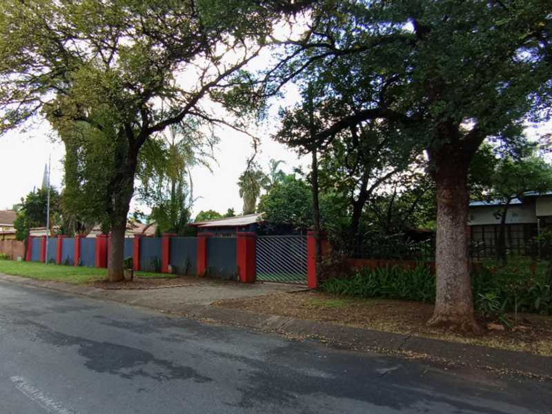 Street view of the property showing the perimeter wall, metal driveway gate, and mature trees along the front boundary.