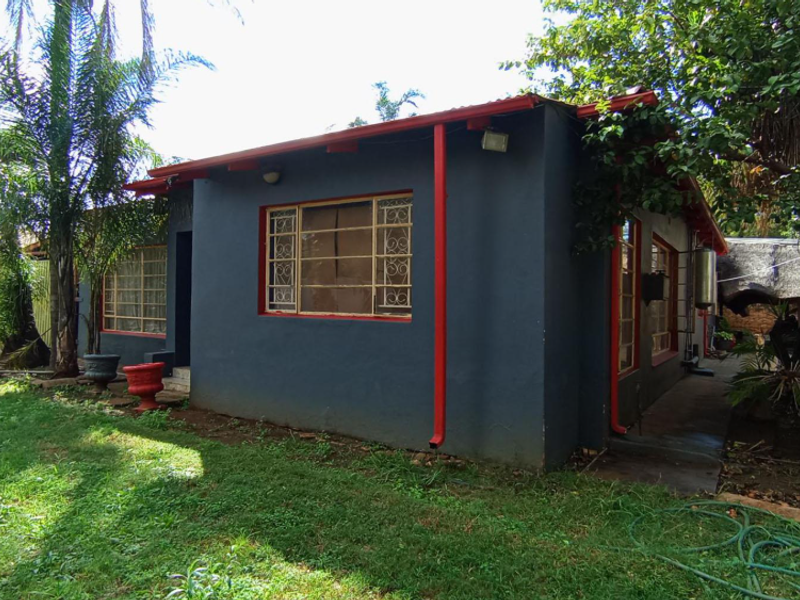 Rear view of a residential house featuring dark grey exterior walls with red trim, metal security window bars, and a surrounding garden area.