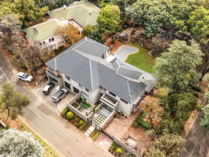Aerial view of a modern residential property featuring a metal roof, paved driveway, swimming pool, and established garden in a leafy suburban setting.