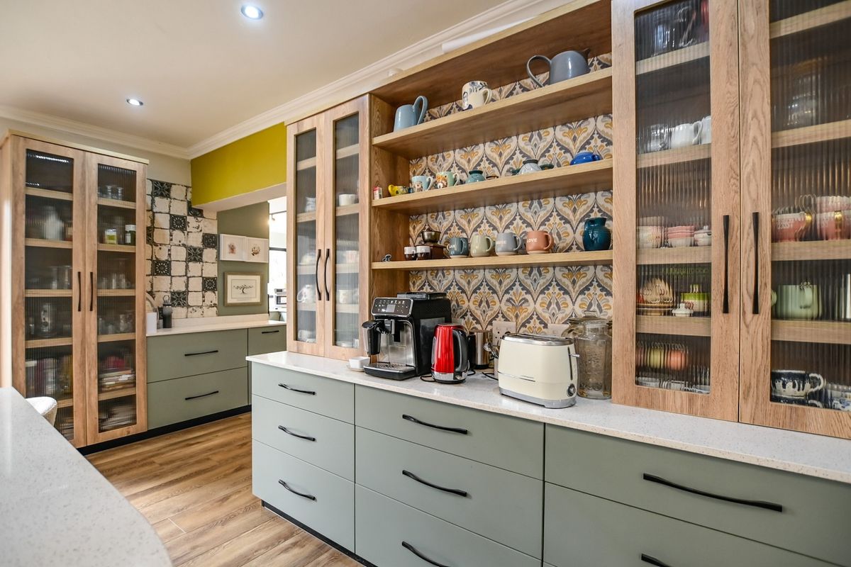 A modern, well-appointed kitchen featuring sage green cabinetry, quartz countertops, fluted glass display cabinets, and decorative patterned tile backsplash.