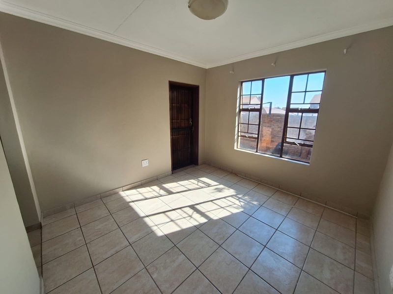 A bright, empty bedroom featuring neutral wall paint, tiled flooring, and a window fitted with security burglar bars.