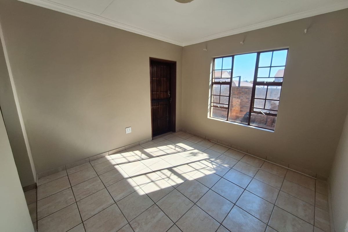 A bright, empty bedroom featuring neutral wall paint, tiled flooring, and a window fitted with security burglar bars.