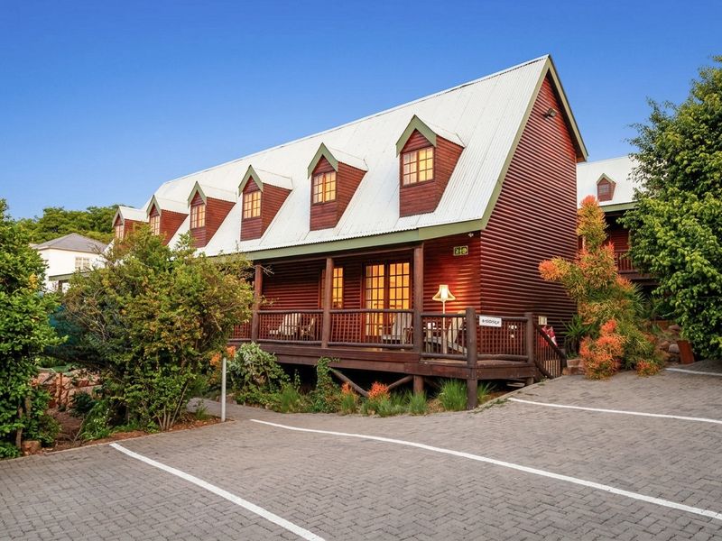 A charming timber-clad house featuring a corrugated metal roof, dormer windows, and a spacious front deck, set above a paved driveway.