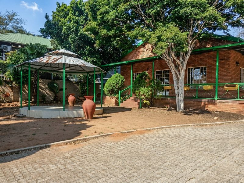 A rear garden area featuring a brick house with a covered patio, a metal gazebo, and a paved driveway surrounded by mature trees.