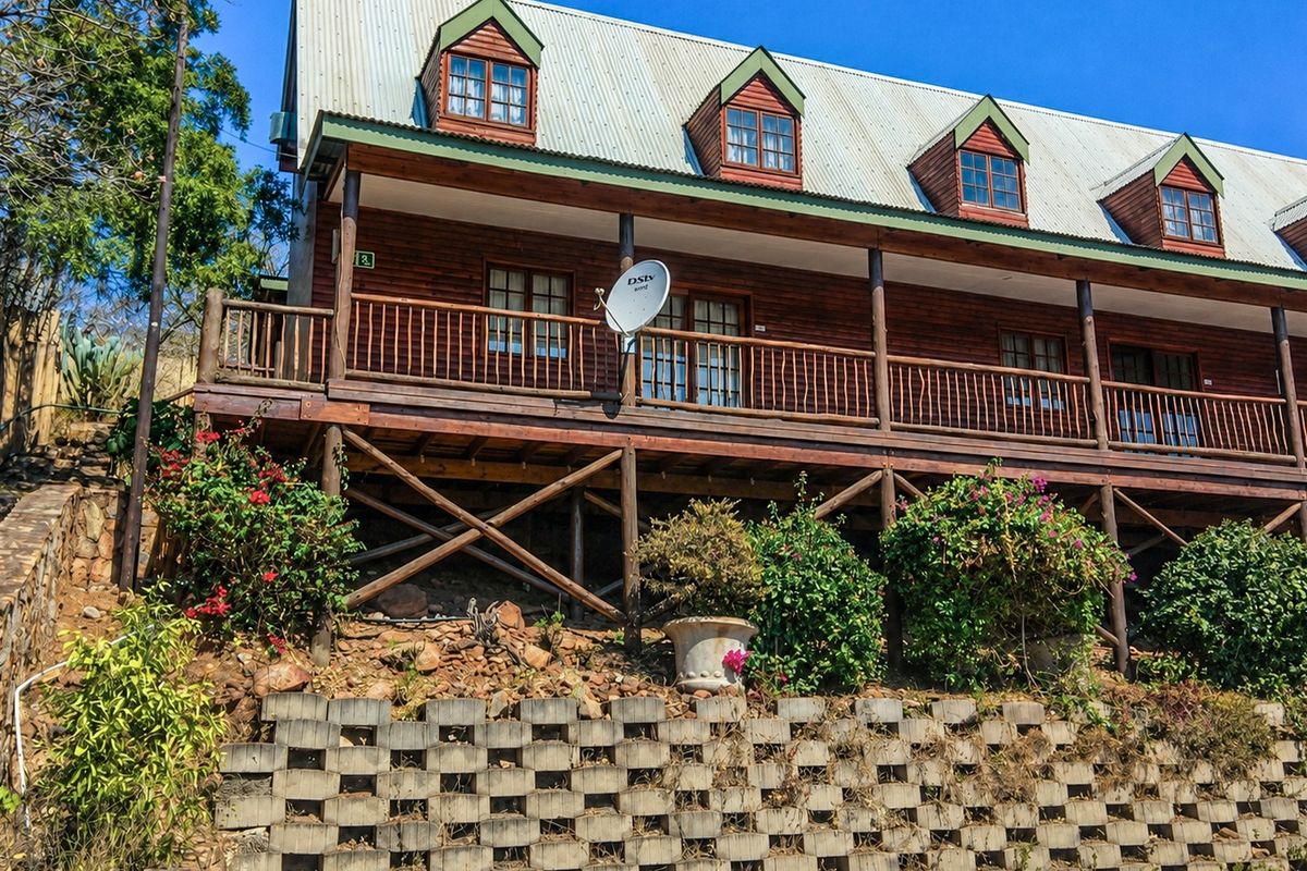 A two-story timber log house featuring a raised wooden deck, dormer windows, and a corrugated metal roof, set above a concrete block retaining wall.