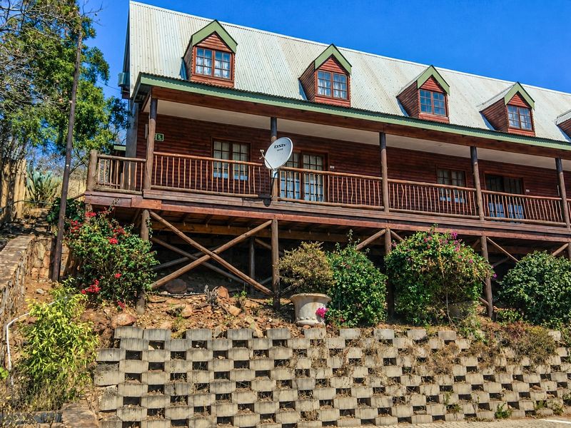A two-story timber log house featuring a raised wooden deck, dormer windows, and a corrugated metal roof, set above a concrete block retaining wall.