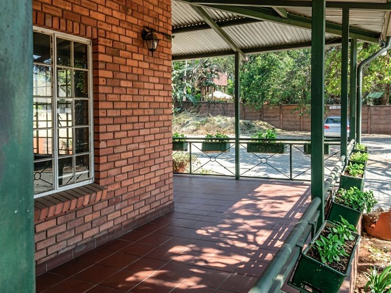 A covered outdoor patio featuring exposed brickwork, tiled flooring, and metal structural supports, overlooking a paved driveway area.
