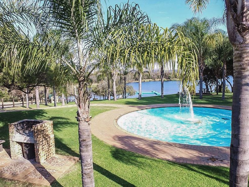 A well-maintained outdoor pool area featuring a circular swimming pool, brick built-in braai, and expansive lawn overlooking a scenic lake.