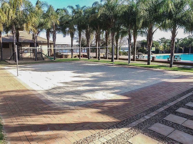 Outdoor recreational area featuring a sand volleyball court, paved pathways, and a swimming pool surrounded by mature palm trees.