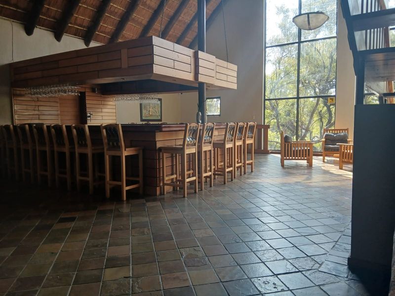Spacious bar area featuring a large built-in wooden counter, suspended canopy, high thatched ceilings, and expansive floor-to-ceiling windows overlooking the surrounding greenery.