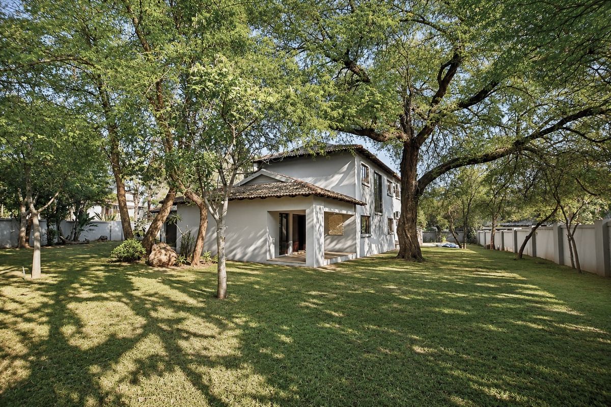 A view of the rear elevation of the house featuring a covered patio, established trees, and a spacious garden enclosed by a perimeter wall.