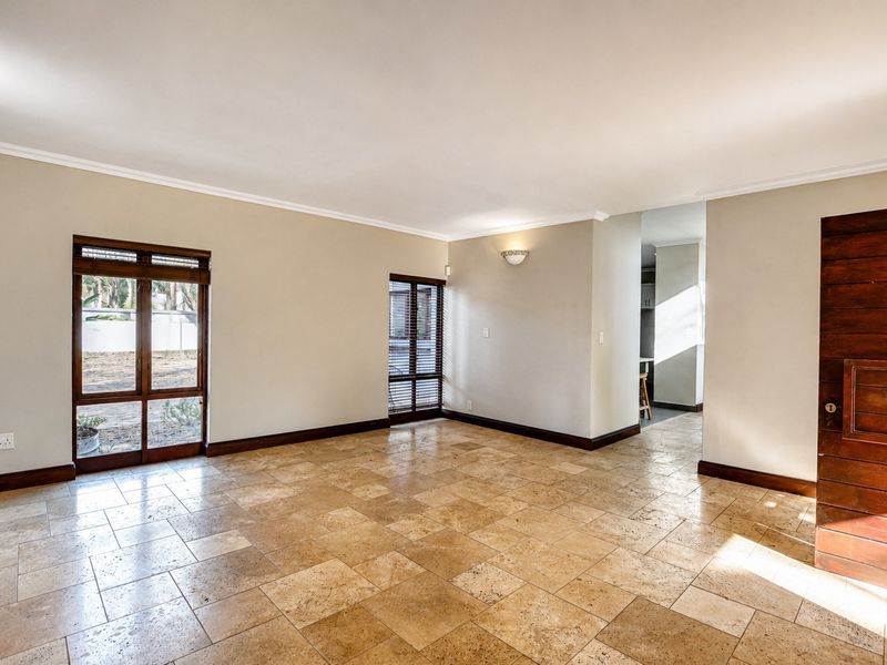 A spacious, empty living area featuring natural travertine floor tiles, wooden window frames, and a solid wooden door, leading through to a kitchen area.