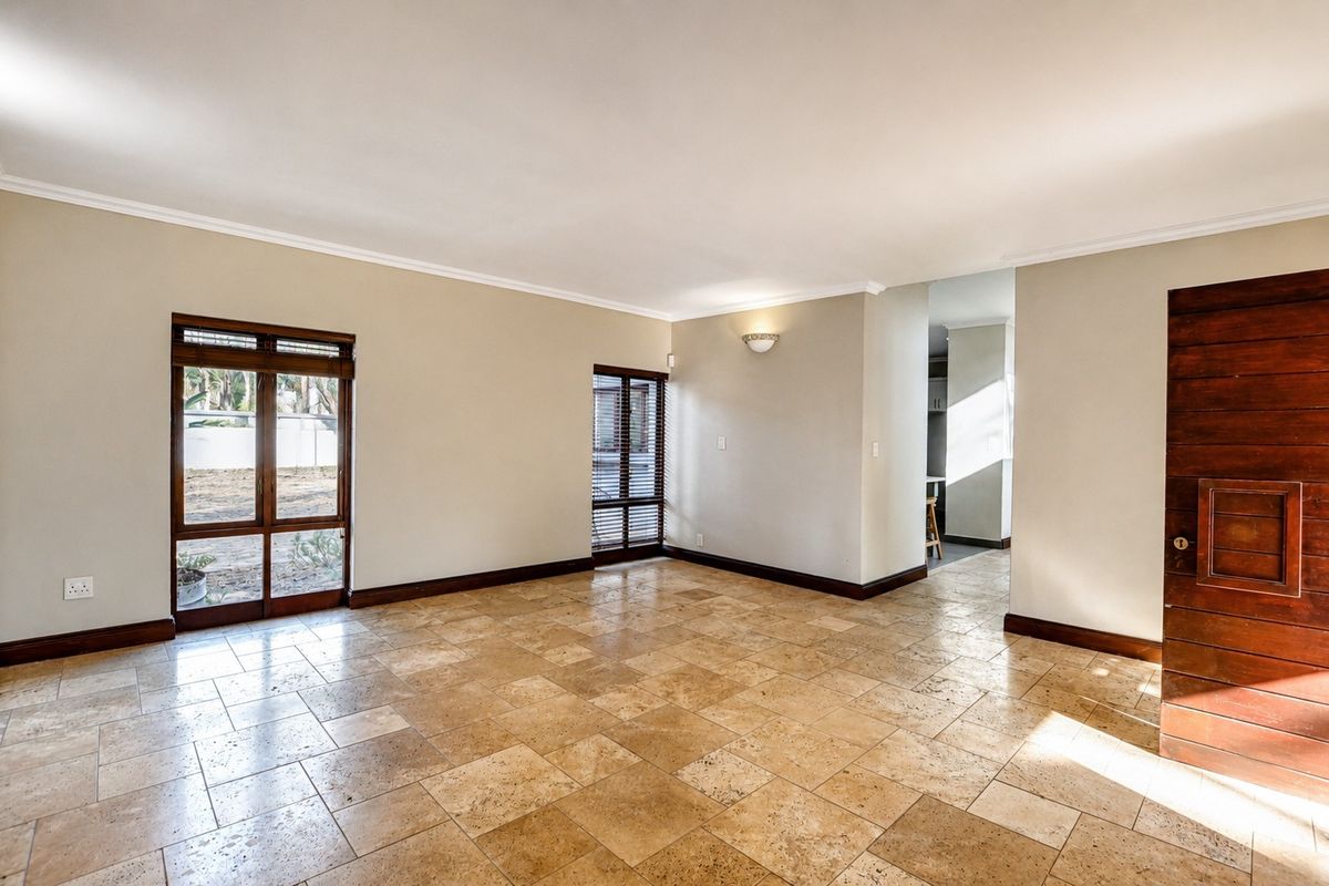 A spacious, empty living area featuring natural travertine floor tiles, wooden window frames, and a solid wooden door, leading through to a kitchen area.