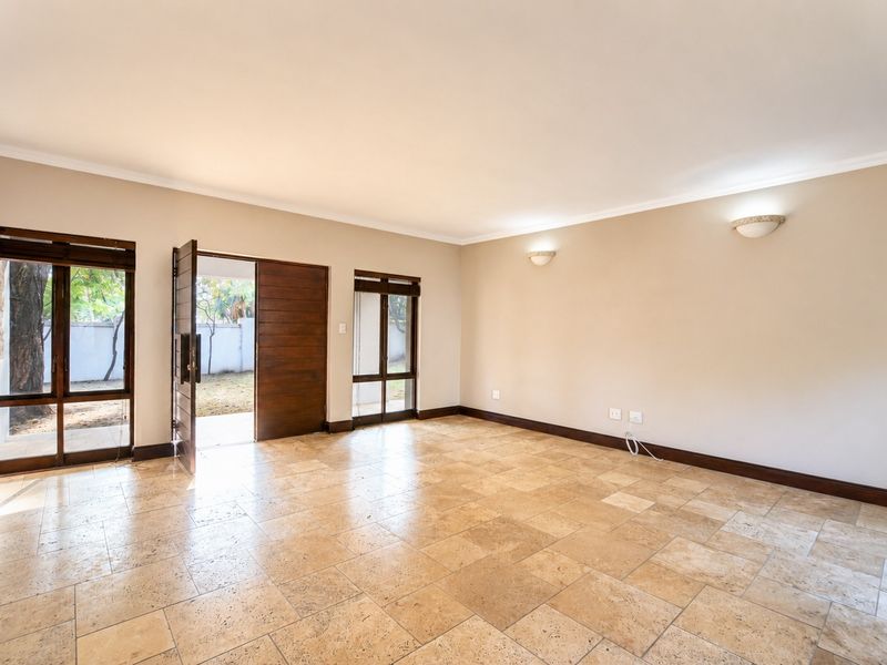 Spacious, empty living area featuring natural travertine tile flooring, timber-framed glass doors leading to the garden, and wall-mounted lighting.