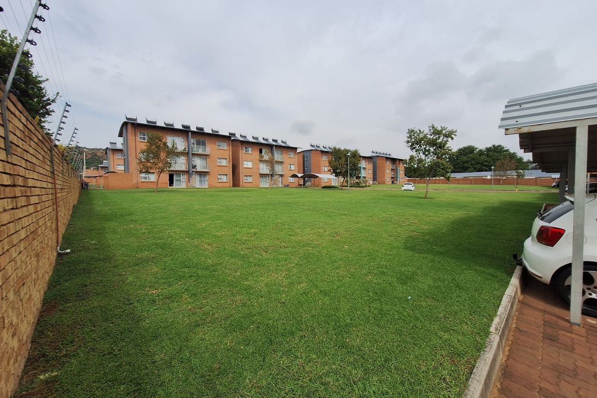 A view of the apartment complex grounds featuring a large communal lawn, brick boundary wall with electric fencing, and covered parking bays.