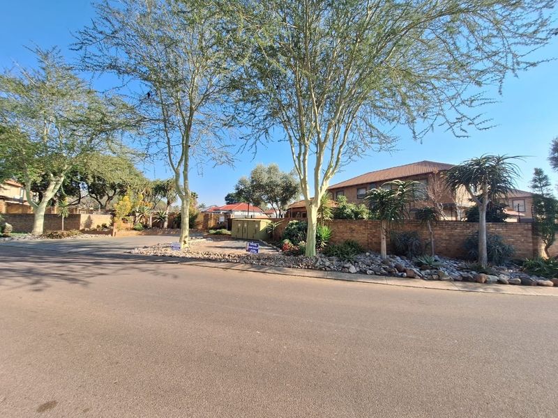 A street-level view of the property exterior, showcasing the brick boundary wall, mature trees, and landscaped verge in a residential townhouse complex setting.