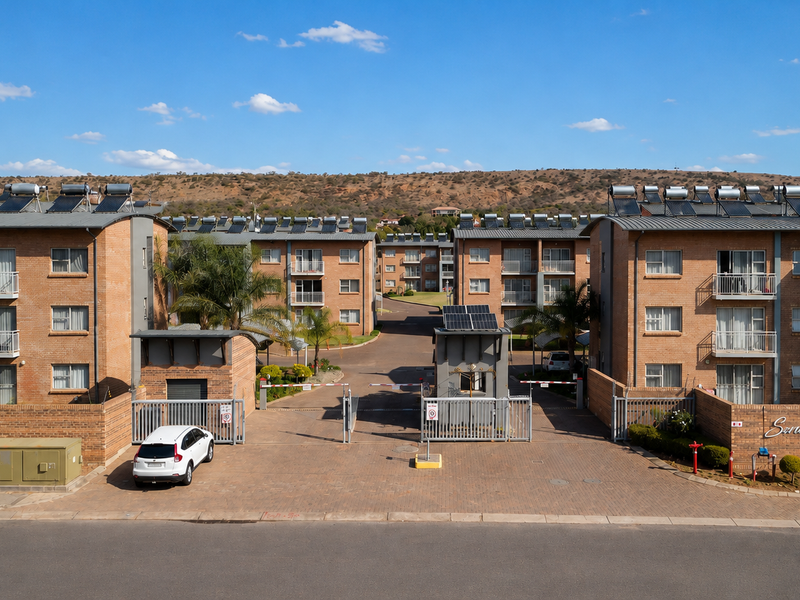 Front view of a multi-story apartment complex featuring brick facades, solar water heating systems, and a secure gated entrance with paved driveway.