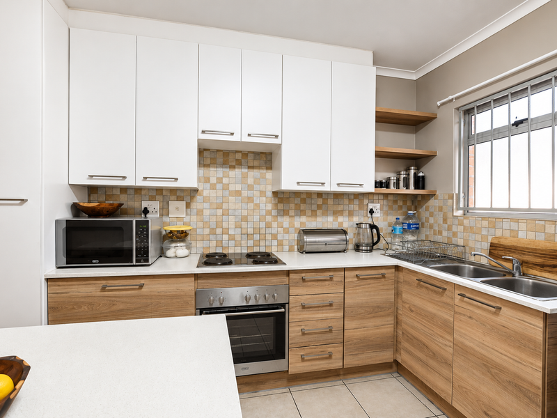 Modern kitchen featuring light wood cabinetry, stone countertops, tiled backsplash, built-in oven, electric hob, and a double sink with security bars on the window.