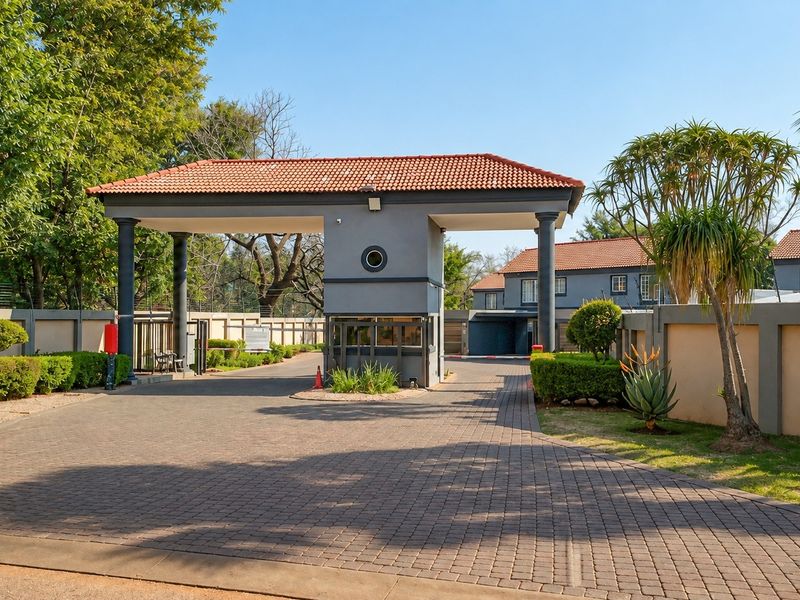 A well-maintained residential complex entrance featuring a secure gatehouse, paved driveway, and perimeter walling with integrated electric fencing.