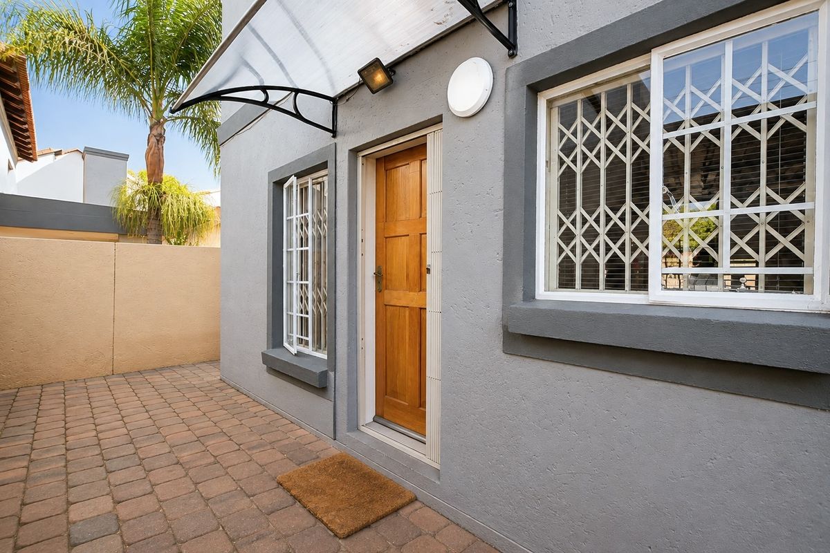 A well-maintained rear exterior area featuring a paved patio, secure entrance door with security gate, and a protective awning over the doorway.